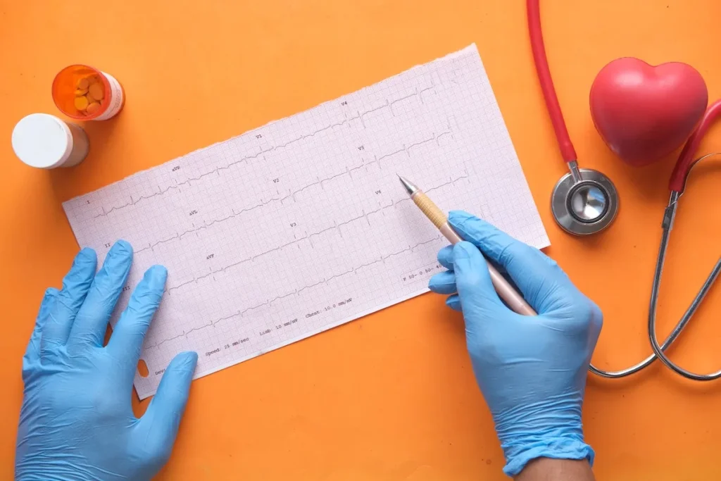A desk surface, with a medical graph, and a medical professional's hands gloved completing the chart with a pencil, with a stethoscope and medicine bottle next to the chart.