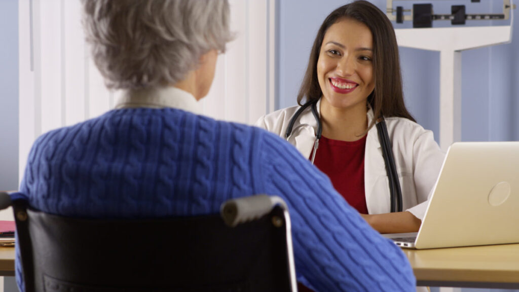 Female doctor with a happy expression and elderly female patient in a wheelchair sitting in an office space with a laptop.