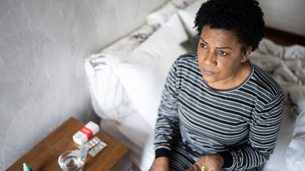 Middle-aged female heart patient with a solemn or sad facial expression, sitting on the edge of a bed, next to a nightstand with medicine bottles. Patient is holding pills in her hand as if she is about to consume them.