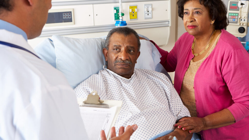 African American male heart patient in the hospital bed next to wife, both looking concerned as they are speaking with a doctor